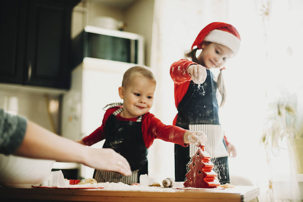 Очаровательная девушка и мальчик в фартуках веселятся за столом с тестом и мукой, делая праздничное печенье с мамой
 - Фото, изображение
