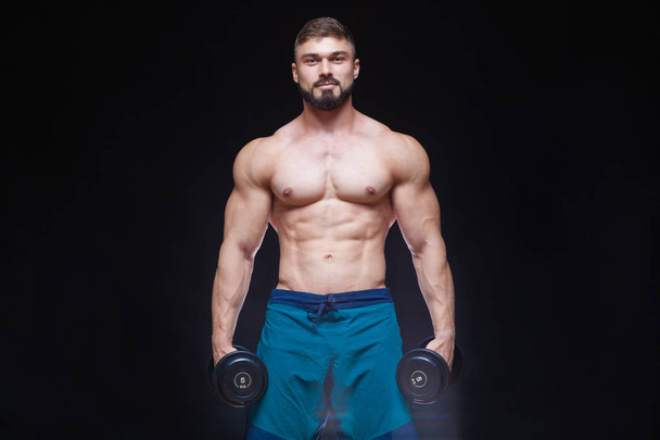 Muscular bodybuilder guy doing exercises with dumbbell over black background - Photo, image
