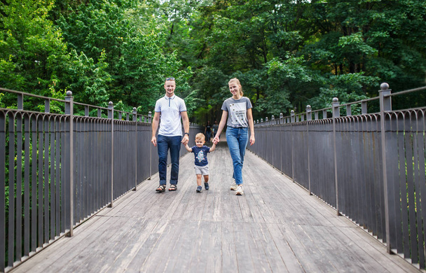 Νέοι χαμογελώντας οικογενειακή εκμετάλλευση χέρια και περπατώντας μέσα από την ξύλινη γέφυρα στο Δημοτικό Πάρκο - Φωτογραφία, εικόνα