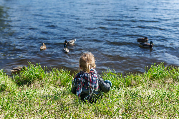 Little girl sitting on grass and looking at lake with floating ducks. Back view - Photo, Image