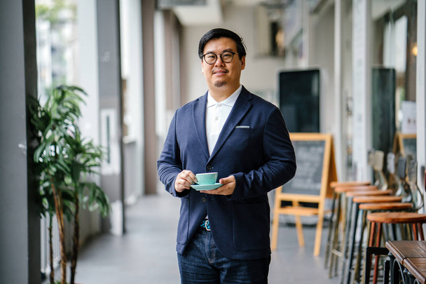 Portrait of an educated, middle-aged Chinese Asian man in business casual and spectacles. He is holding and drinking tea from an elegant cup and saucer during the day in the city. - Foto, Bild