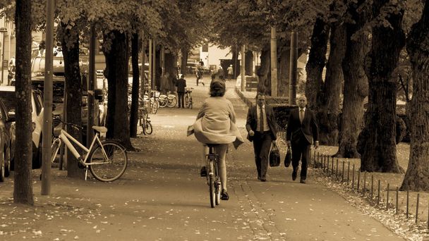 Helsinki, Finlândia - 4 de setembro de 2018. Uma mulher a andar de bicicleta de gabardina, calças cor-de-rosa e um capacete de protecção no parque. A caminho de dois homens em ternos de negócios
 - Foto, Imagem