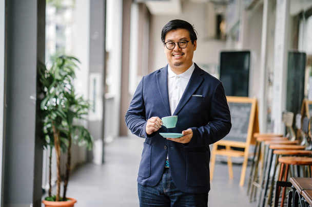 Portrait of an educated, middle-aged Chinese Asian man in business casual and spectacles. He is holding and drinking tea from an elegant cup and saucer during the day in the city. - Фото, изображение