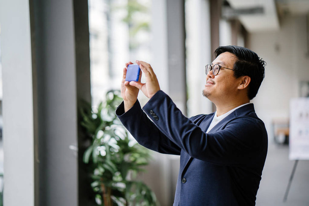 Portrait of a confident middle-aged Chinese Asian man taking a photograph on his smartphone during the day. He is dressed in smart casual (polo, jacket and jeans). - 写真・画像