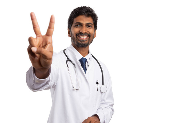 Hymyilevä intialainen mies lääkintämies, joka näyttää rauhan tai voiton merkkiä etusormella ja keskisormella eristettynä valkoisella studion taustalla
 - Valokuva, kuva