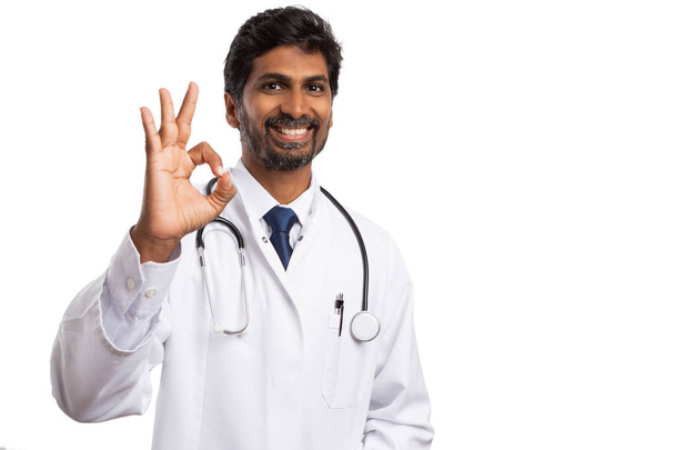 Индийский врач улыбается и держит пальцы, как хорошо знак изолирован на белом фоне студии
 - Фото, изображение