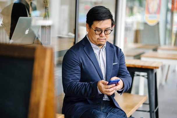 Portrait of a middle-aged Chinese Asian man in a casual suit (jacket, oxford button-down shirt and jeans) and glasses using his smartphone as he sits on a bench outside a cafe in the city. - Fotó, kép