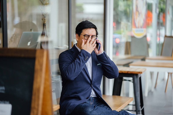 Portrait of a middle-aged Chinese Asian man in a casual suit (jacket, oxford button-down shirt and jeans) and glasses talking on his smartphone as he sits on a bench outside a cafe in the city. - Foto, Imagen