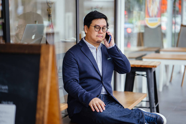 Portrait of a middle-aged Chinese Asian man in a casual suit (jacket, oxford button-down shirt and jeans) and glasses talking on his smartphone as he sits on a bench outside a cafe in the city. - Fotoğraf, Görsel