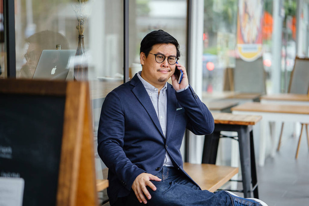 Portrait of a middle-aged Chinese Asian man in a casual suit (jacket, oxford button-down shirt and jeans) and glasses talking on his smartphone as he sits on a bench outside a cafe in the city. - Фото, изображение