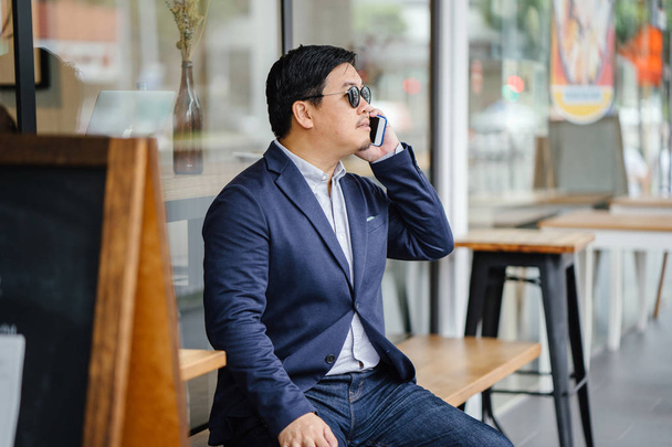 Portrait of a middle-aged Chinese Asian man in a casual suit (jacket, oxford button-down shirt and jeans) and sunglasses talking on his smartphone as he sits on a bench outside a cafe in the city. - Фото, зображення