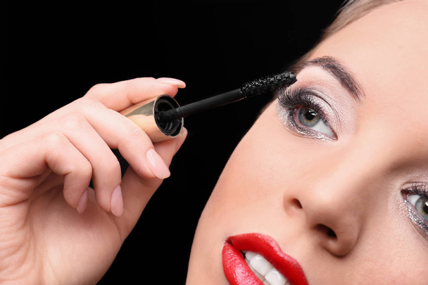 Beautiful young woman with eyelash extensions applying mascara on black background, closeup - Zdjęcie, obraz
