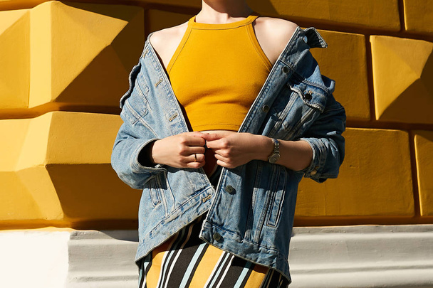 A woman with bare shoulders in a denim blue jacket and in a yellow top on a yellow textured wall. Street style. - Фото, изображение
