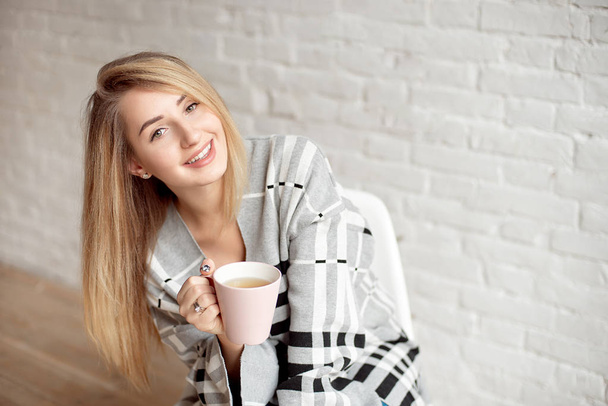 Young happy girl wrapped in a warm blanket looks happy holding cup of tea indoors - Photo, Image