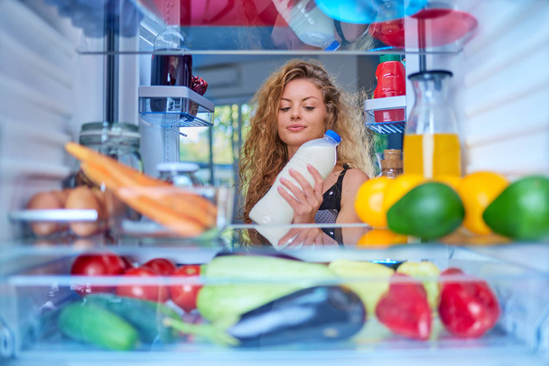 Woman standing in front of fridge and taking milk. Fridge full of groceries. - Photo, Image