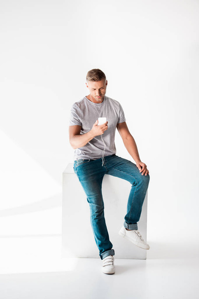 handsome adult man in blank grey t-shirt listening music with smartphone and earphones on white - Foto, Imagem