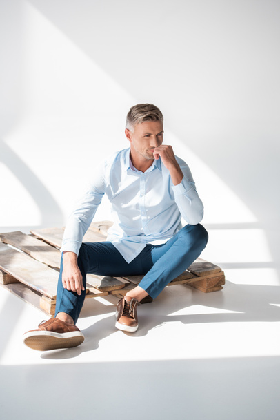 ελκυστική ενηλίκων άνθρωπος σε μοντέρνα ρούχα κάθεται σε ξύλινη παλέτα σε λευκό και αναζητούν μακριά - Φωτογραφία, εικόνα