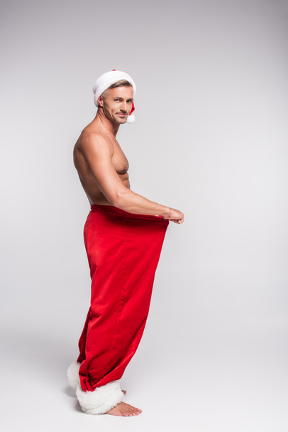 side view of shirtless man in santa hat holding red pants and smiling at camera on grey  - Фото, зображення