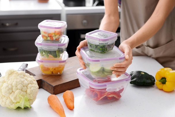 Woman holding stack of plastic containers with fresh vegetables for freezing at table in kitchen - Photo, Image