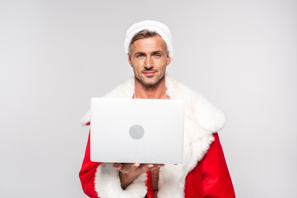 handsome man in santa costume holding laptop and smiling at camera isolated on grey   - Zdjęcie, obraz