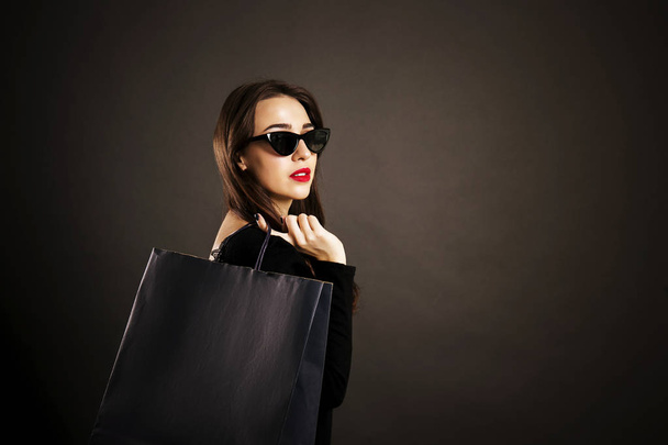 Black friday sale concept. Attractive young woman with long brunette hair, smiling, wearing sexy dress & cat eye sunglasses, holding blank shopping bags over black background. Copy space, close up. - Photo, Image