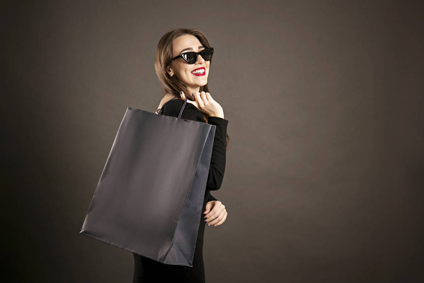 Black friday sale concept. Attractive young woman with long brunette hair, smiling, wearing sexy dress & cat eye sunglasses, holding blank shopping bags over black background. Copy space, close up. - Foto, afbeelding