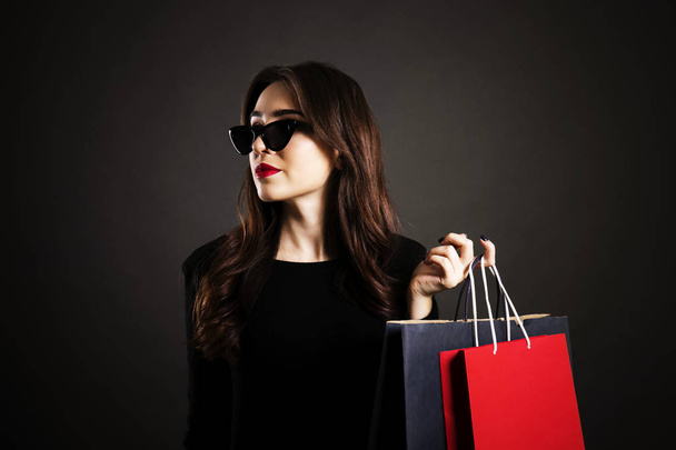 Black friday sale concept. Attractive young woman with long brunette hair, smiling, wearing sexy dress & cat eye sunglasses, holding blank shopping bags over black background. Copy space, close up. - Zdjęcie, obraz