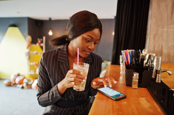 Afrikkalainen liikenainen juo cocktaillimonadia kahvilassa baarissa. Musta tyttö ottaa levätä matkapuhelimella
. - Valokuva, kuva