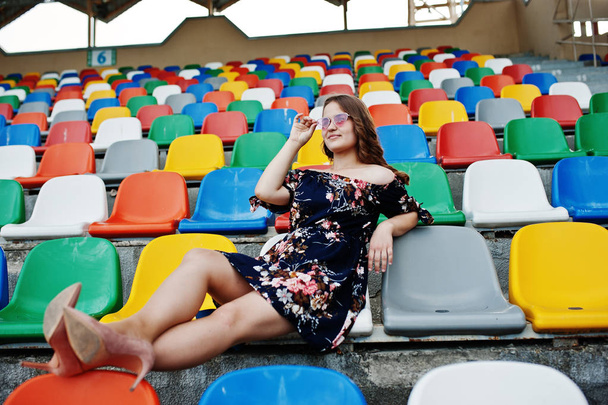 Portrait of a young beutiful girl in dress and sunglasses sitting on the tribunes in stadium. - Foto, immagini