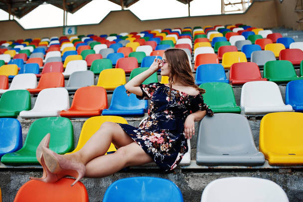 Portrait of a young beutiful girl in dress and sunglasses sitting on the tribunes in stadium. - 写真・画像