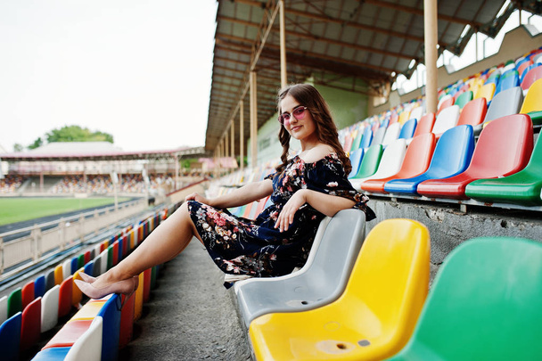 Portrait of a young beutiful girl in dress and sunglasses sitting on the tribunes in stadium. - Фото, зображення