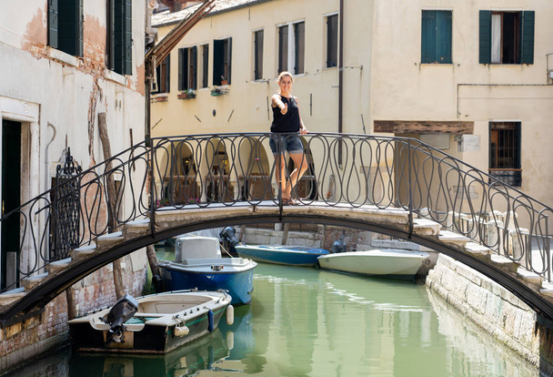 Atractiva turista latina natural posando y mirando la cámara mostrando el pulgar hacia arriba en un puente en el canal en Venecia En Vacaciones de Verano Viajando por Europa concepto
. - Foto, imagen