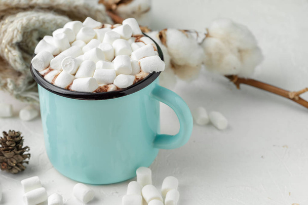 Blue metal mug of cocoa or hot chocolate with marshmallows on the table with winter decor, branch of corton flower, knitted blanket and wooden Christmas ornaments - Fotoğraf, Görsel