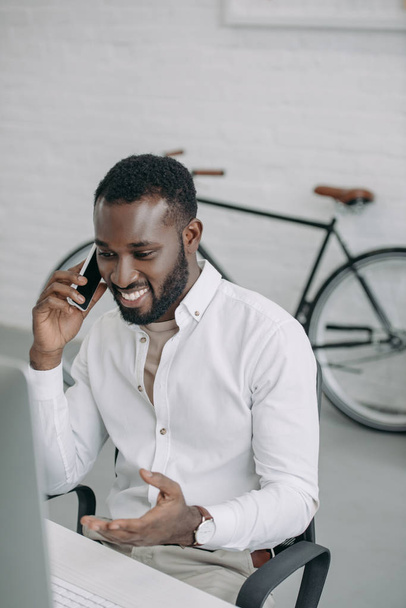 smiling handsome african american businessman talking by smartphone and gesturing in office - Fotó, kép
