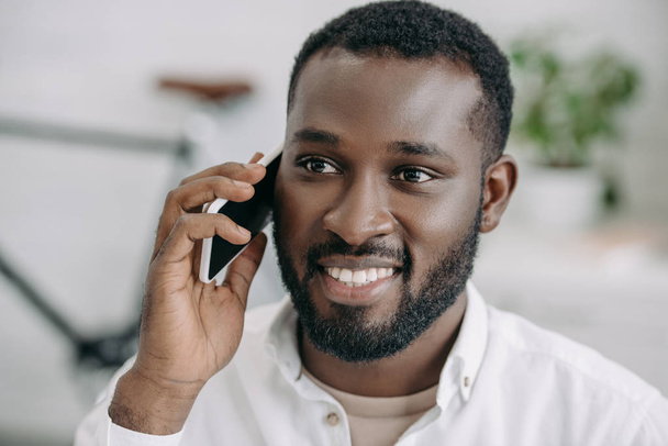 portrait of smiling handsome african american businessman talking by smartphone in office - Fotografie, Obrázek
