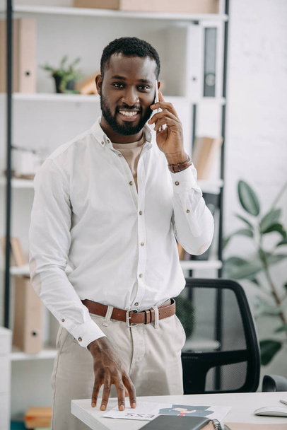handsome african american businessman standing near table and talking by smartphone in office - Фото, зображення
