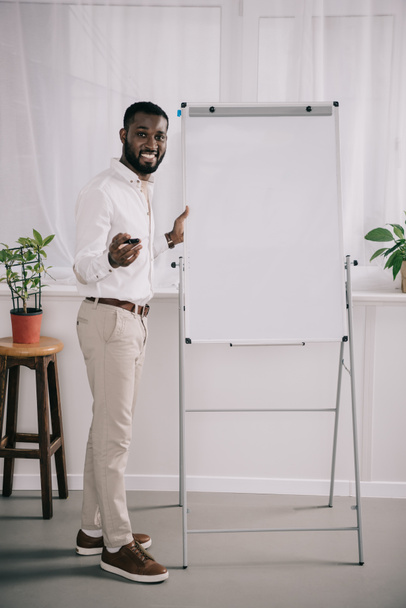 улыбающийся красивый африканский бизнесмен, стоящий рядом с флипчартом и жестикулирующий в офисе
 - Фото, изображение