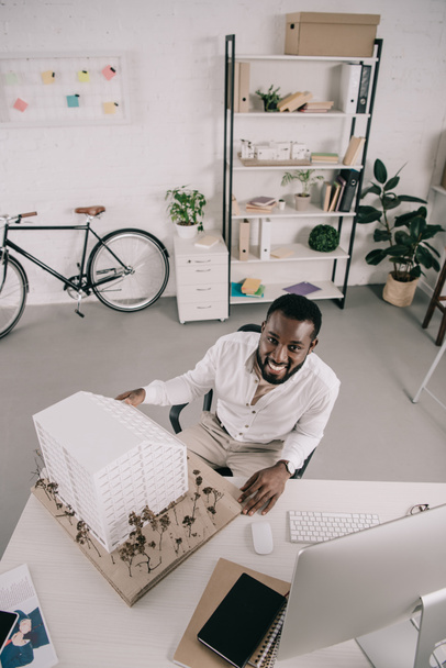 hoge hoekmening van lachende knappe Afro-Amerikaanse architect zit in de buurt van het platform model en kijken naar camera in kantoor - Foto, afbeelding