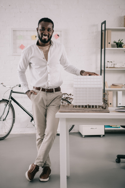 усміхнений красивий афроамериканський архітектор торкається архітектурної моделі і дивиться на камеру в офісі
 - Фото, зображення