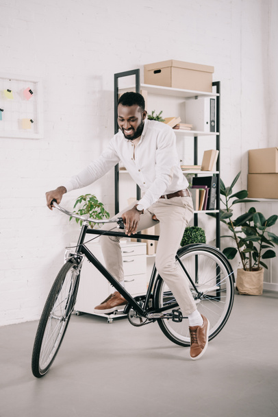 smiling handsome african american businessman riding bike in office - Foto, immagini