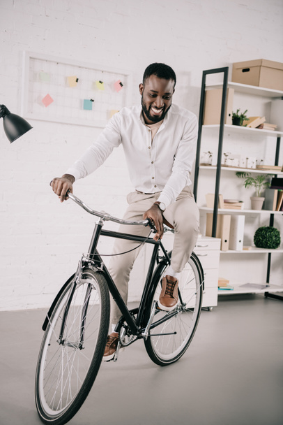 smiling handsome african american businessman riding bicycle in office - Foto, immagini