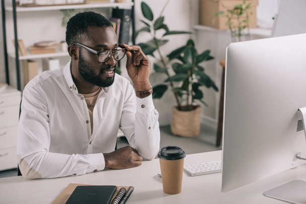 χαμογελώντας όμορφος Αφρικανικός Αμερικανός επιχειρηματίας αγγίζοντας γυαλιά και να βλέπουν τον υπολογιστή στο γραφείο - Φωτογραφία, εικόνα