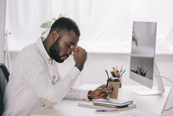 вид збоку втомлений афроамериканський бізнесмен торкається моста носа за столом з комп'ютером в офісі
 - Фото, зображення