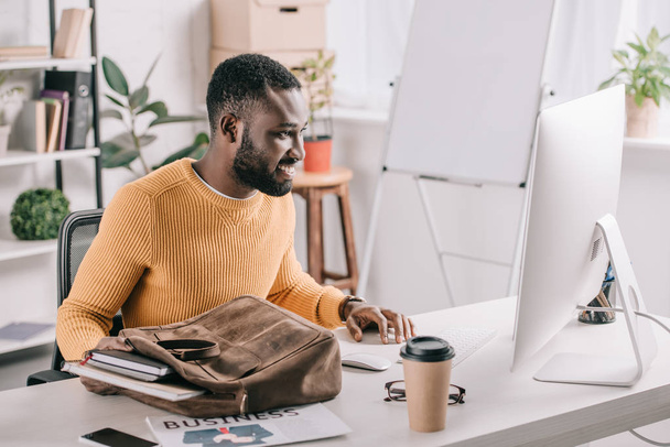 handsome african american designer in orange sweater taking notebooks from briefcase and looking at computer in office - Foto, imagen