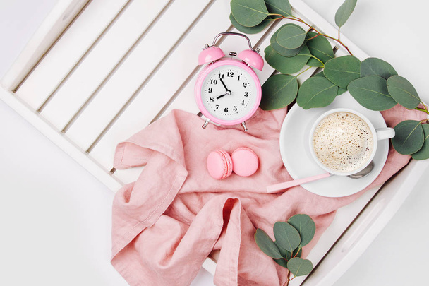 Cup of coffee with macarons, clock and eucalyptus branches on serving tray with pink fabric - Foto, Imagem
