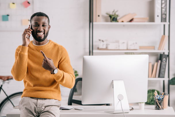 uśmiechający się przystojny Afryki amerykański projektant w pomarańczowy sweter rozmowy za pomocą smartfonów i wskazując na gadżet w biurze - Zdjęcie, obraz