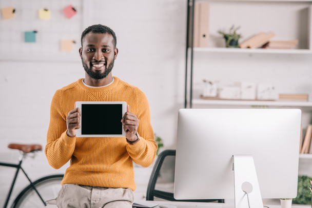 улыбающийся красивый африканский американский дизайнер в оранжевом свитере показывает планшет с пустым экраном в офисе
 - Фото, изображение