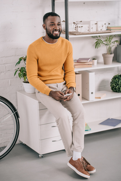 handsome cheerful african american designer in yellow sweater using smartphone in office - Zdjęcie, obraz