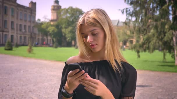 Good-looking caucasian short-haired blonde female is scrolling her phone while standing outside, near old city and park - Кадри, відео