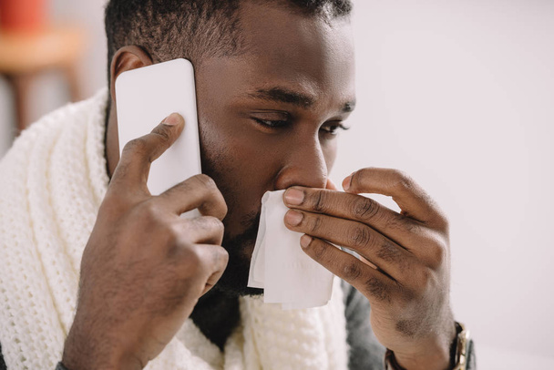 hombre afroamericano enfermo con nariz líquida sosteniendo la servilleta mientras habla en el teléfono inteligente
 - Foto, Imagen